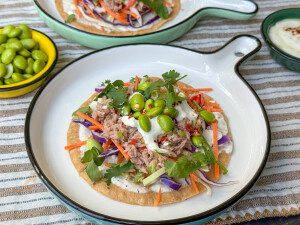 tostada met tonijn en avocado, sojaboontjes en koolmix geserveerd met een frisse limoen – yoghurtdipsaus geserveerd op een bord.