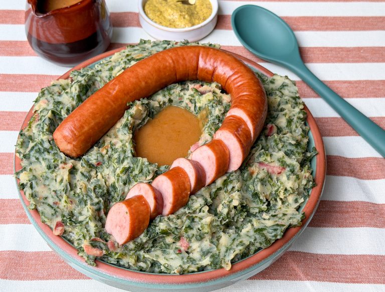 Boerenkool stamppot met rookworst, gebakken spekjes en grove mosterd geserveerd in schaal op tafel.