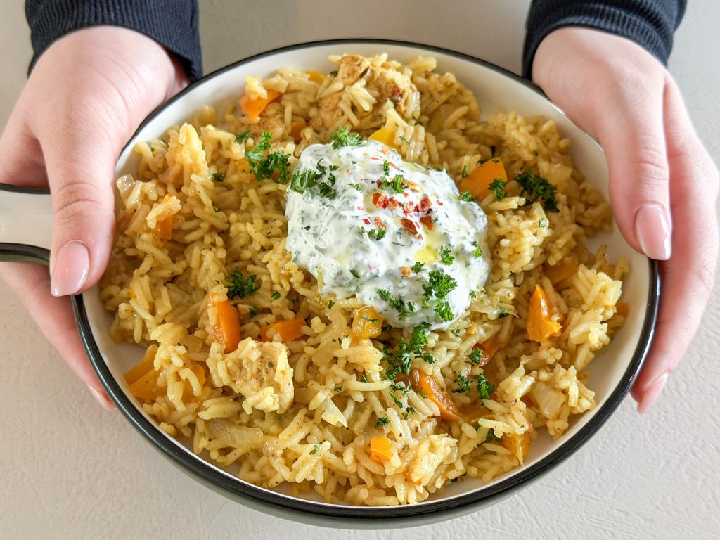 Close-up van een bord met eenpansgerecht met rijst, kip en yoghurtdip in twee handen vastgehouden