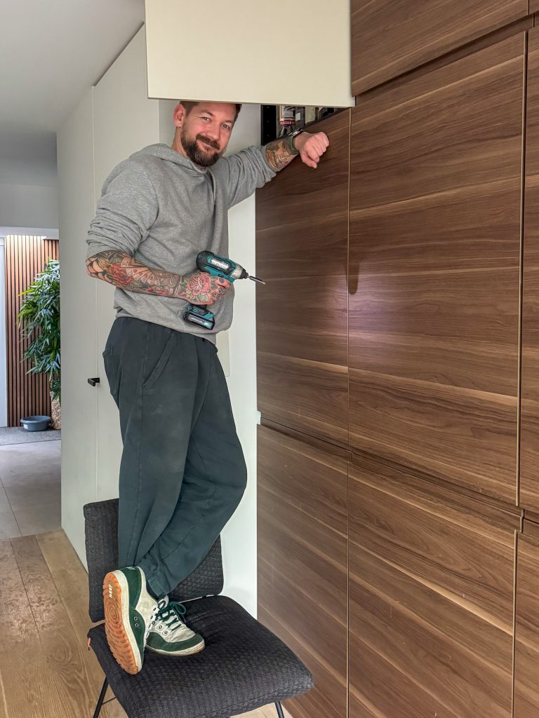 Jan Willem staat op een stoel en werkt met de boormachine aan de hoge kast.