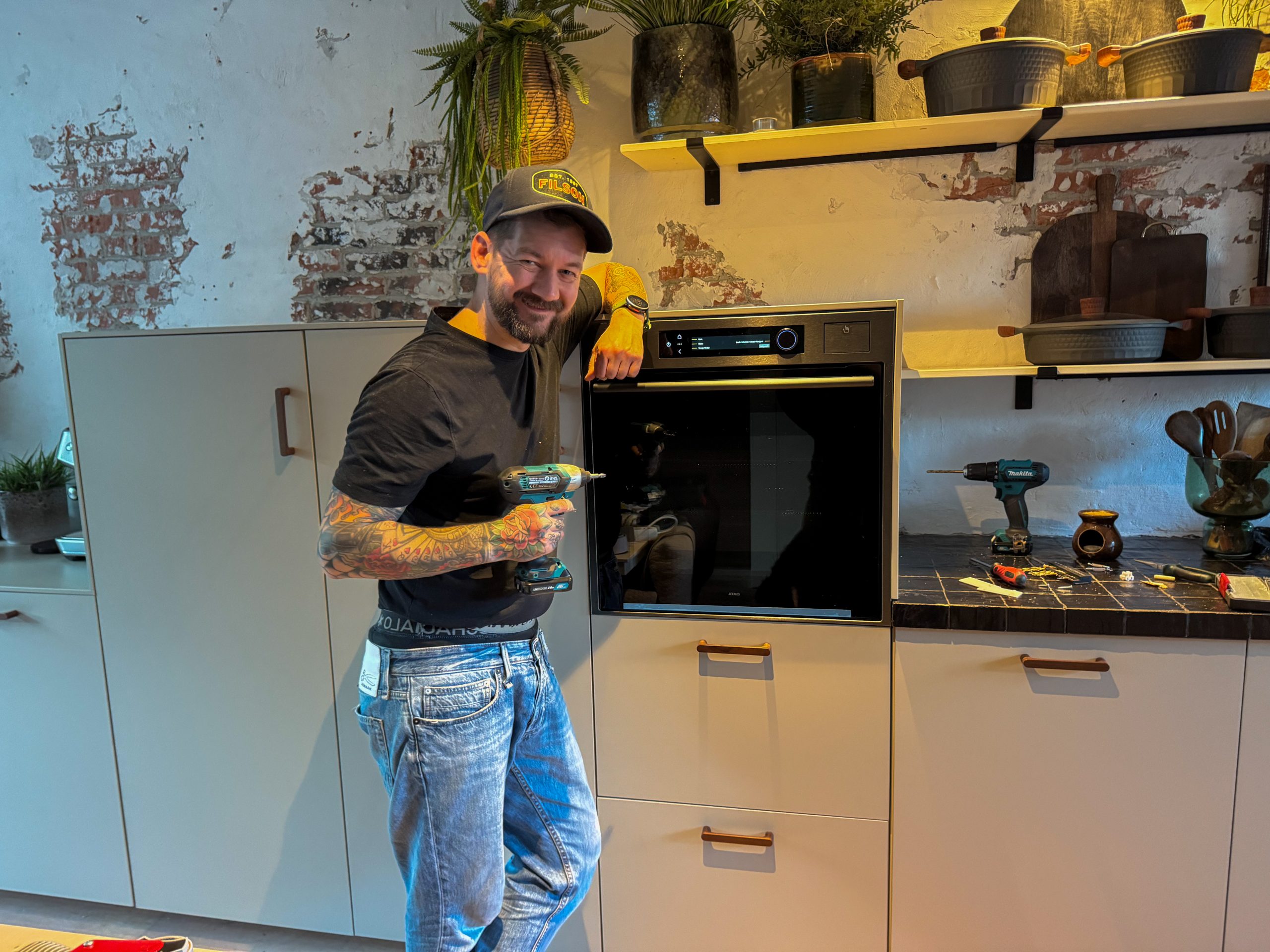 Jan Willem plaatst de nieuwe oven in de keuken en houdt een boormachine vast.